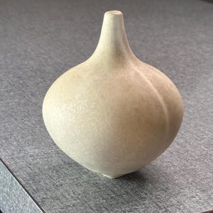 以下が含まれることがあります： 狭い首と丸みを帯びたタマネギのような形状の胴体を持つ、小さな球根状の陶器製花瓶。表面はマットなオフホワイトの仕上げで、全体に淡いベージュとクリーム色の斑点状の質感が施されています。