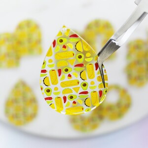 May include: A clear teardrop-shaped resin pendant with a colourful pattern of yellow, red, and green tacos, avocados, and limes.