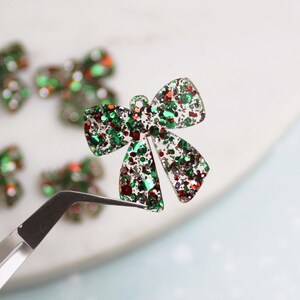 May include: A close-up of a green and red glitter bow charm. The bow is made of clear plastic and is filled with green, red, and silver glitter. The bow is being held by a pair of tweezers.