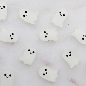 May include: A collection of small, white ghost-shaped embellishments with black smiling faces. These Halloween-themed decorations are scattered across a light-colored surface, creating a playful and spooky aesthetic.