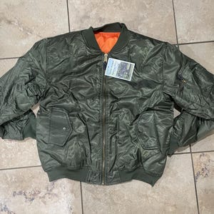 May include: A green bomber jacket with a zip fastening and a contrasting orange lining. The jacket has a pocket on the left side and a tag attached to the zip.