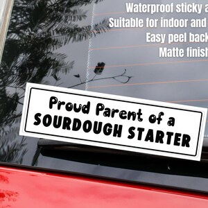 May include: A white bumper sticker on a red car window with the text "Proud Parent of a Sourdough Starter." The sticker is rectangular with a black border. Additional text describes the sticker's material and features.