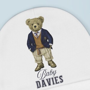 May include: A white baby beanie featuring a charming polo bear design. The bear is wearing a navy blazer, khaki pants, and brown shoes. The text "Baby DAVIES" is printed on the beanie. A yellow star-shaped toy is in the background.
