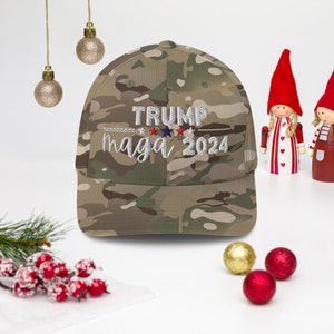 Puede incluir: Una gorra de b&eacute;isbol de camuflaje con el texto bordado "TRUMP maga 2024" en blanco. La gorra presenta estrellas rojas, blancas y azules. El sombrero es una mezcla de colores verdes, marrones y beige.