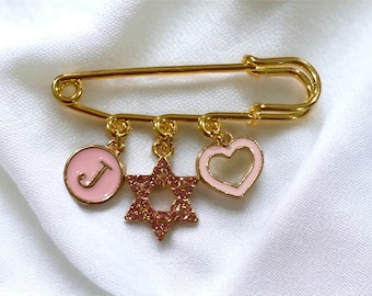 May include: Gold safety pin with three charms: a pink circle with the letter J, a pink star with rhinestones, and a pink heart with a cutout.