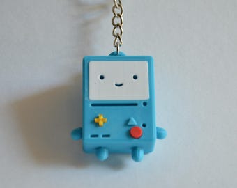 May include: A light blue rectangular keychain featuring a white screen face with two small dots for eyes and a curved line for a mouth. The body includes a yellow plus-shaped button, a blue triangle, and a red circular button on the front surface.