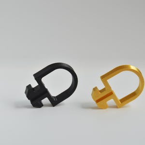 May include: Two D-shaped plastic clips, one in matte black and one in metallic gold. Each clip features a rounded loop on one side and a notched, rectangular attachment point on the opposite side, designed for securing or organizing items.