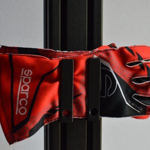 Op de afbeelding: Een rood-zwarte Sparco K-Attack racehandschoen vastgehouden door een zwarte metalen montagebeugel. De handschoen is voorzien van witte Sparco-branding op de pols en zwarte geometrische patronen op de stof, met zichtbare stiksels bij de vingers.
