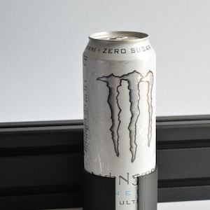 May include: A white Monster Energy Ultra drink can with a silver claw logo and the text "TAURINE + ZERO SUGAR" printed near the top. The can is held in a black cylindrical plastic holder with a vertical cutout, positioned against a black rail.