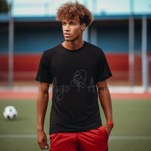 Könnte beinhalten: Ein Mann trägt ein schwarzes T-Shirt mit einem einfachen Strichzeichnungen-Design auf der Vorderseite. Das Shirt hat kurze Ärmel und einen Rundhalsausschnitt. Der Mann steht auf einem grünen Feld mit einem Fußball im Hintergrund. Der Text "Indigo Arts" ist auf dem Shirt sichtbar.