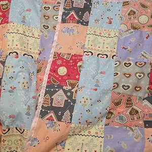 May include: A patchwork quilt with various fabric patterns. The quilt features squares with gingerbread houses, trees, hearts, and figures in shades of blue, pink, red, and grey. A pink satin ribbon borders the quilt.