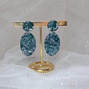 Peut inclure: Boucles d'oreilles pendantes avec un motif marbré. Elles présentent un motif tourbillonnant vert foncé et lavande avec des accents argentés. Chaque boucle d'oreille a un petit dessus circulaire et un fond ovale. Les boucles sont exposées sur un présentoir doré.
