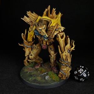 May include: A painted miniature of a wooden creature with a yellow cloth draped over its body. The creature has a human-like face and is standing on a circular base with a black and white dice.