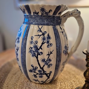 May include: A white and blue ceramic jug with a floral pattern. The jug has a detailed blue floral design on a white background, with a decorative handle and rim. The jug is a decorative item.