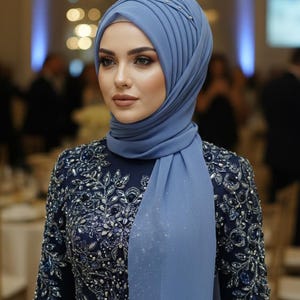 May include: A woman in a blue hijab and a navy dress with silver embroidery. The dress features long sleeves and a detailed floral design. The hijab is a lighter blue and has small decorative elements.