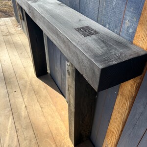 May include: A black wooden mantel shelf with two black wooden legs. The shelf is mounted to a wall with wood paneling.