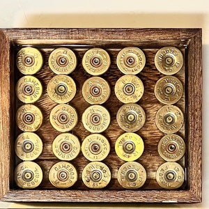 May include: A wooden frame with a glass front displays a collection of 25 gold shotgun shells. The shells are arranged in a grid pattern and are all the same size and shape.