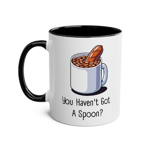 May include: White ceramic mug with a black rim and handle. The mug has a cartoon illustration of a mug filled with beans and a sausage. The text "You Haven't Got A Spoon?" is printed below the illustration.