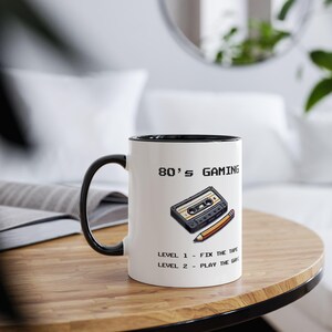 May include: White ceramic mug with a black rim and handle. The mug has a graphic of a cassette tape and pencil with the text "80's GAMING" and "LEVEL 1 - FIX THE TAPE" and "LEVEL 2 - PLAY THE GAME".
