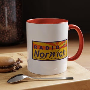 May include: White ceramic mug with a red rim and handle. The mug features a yellow and blue logo that says "RADIO NORWICH".