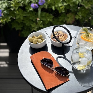 May include: A brown leather glasses case with a metal ring on a gray table. There are two glasses, a bowl of olives, and a bowl of nuts on the table.