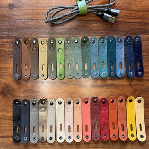 Op de afbeelding: Echte leren kabelbinders in verschillende kleuren. De afbeelding toont een selectie van kabelbinders in verschillende tinten, van bruin en groen tot blauw en roze. De tekst "30 kleuren beschikbaar" is ook zichtbaar.