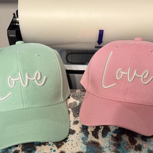 May include: Two baseball caps, one green and one pink, with the word "Love" embroidered in white on the front of each.