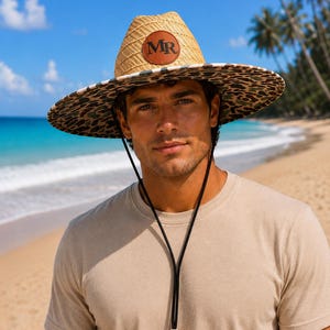 May include: A straw hat with a wide brim featuring a leopard print pattern on the underside. The crown is made of woven straw and includes a circular brown leather patch with the letters MR embossed in the center. A black chin cord is attached.