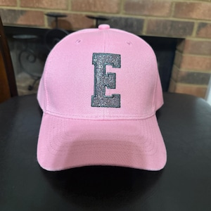 Puede incluir: Gorra de béisbol rosa con visera curva. La gorra presenta una gran letra "E" brillante en la parte delantera. El sombrero está hecho de una tela suave y tiene un botón en la parte superior.