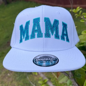 Könnte beinhalten: Eine weiße Snapback-Sportkappe mit dem Wort "MAMA" in türkisfarbenen Blockbuchstaben gestickt. Die Kappe hat einen gebogenen Schirm und einen Schnappverschluss. Ein "SNAPBACK sport cap"-Etikett befindet sich auf der Vorderseite.