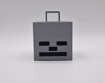 May include: A grey, cube-shaped object with a handle on top and a black skull face design. The face features two rectangular eye shapes and a horizontal mouth shape. The object is likely a decorative item or toy.