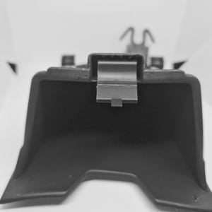 Op de afbeelding: Een zwart kunststof auto-onderdeel, mogelijk een sierlijst of beugel, met een metalen clip. Het onderdeel heeft een geometrische vorm met een gebogen onderrand en een rechthoekige clip in het midden. De achtergrond is wit.