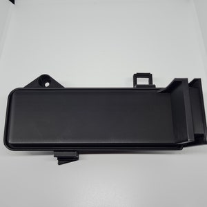 Puede incluir: Pieza de automóvil de plástico negro con forma rectangular y múltiples pestañas y clips.
