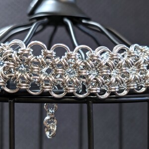 Peut inclure: Un bracelet en cotte de mailles argenté avec un motif floral. Le bracelet est composé de petits anneaux métalliques entrelacés. Une petite chaîne pend du bas. Le bracelet est présenté sur un support en métal noir.