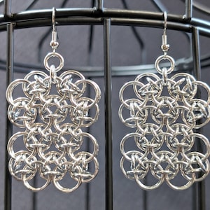 Peut inclure: Une paire de boucles d'oreilles en cotte de mailles argentées. Chaque boucle d'oreille présente un motif rectangulaire composé d'anneaux métalliques entrelacés. Les boucles d'oreilles sont suspendues à un crochet simple. Le fond est gris foncé.