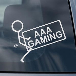 May include: White vinyl car decal featuring a stick figure carrying a sign that reads "AAA GAMING." The decal is displayed on a car's rear window. The text "Example display only - see dimensions in listing" is at the bottom.