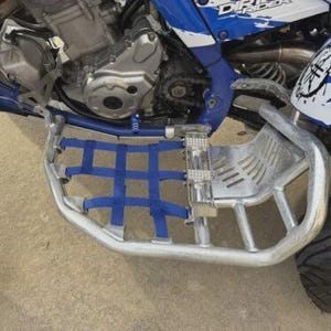 Może przedstawiać: Zbliżenie podnóżka ATV z niebieską siatką i srebrną metalową ramą. ATV ma niebieskie i białe akcenty. Opona jest czarna, a podłoże brązowe.