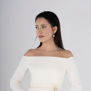 Puede incluir: Vestido marfil con hombros descubiertos y mangas largas. El vestido presenta un cinturón de cuentas con un adorno de plumas y joyas. La modelo lleva pendientes de perlas. Diseño clásico y elegante.
