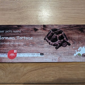 May include: A wooden sign featuring a brown tortoise. The sign reads "Your pet's name: Herman's Tortoise, Testudo Hermanni". It also includes a map of Europe with a green area indicating the tortoise's native range. The sign has a red circle with the words "Least Concern" on it.