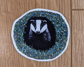 May include: A round sticker with a white border featuring a black and white badger peeking out of a burrow surrounded by green grass and blue flowers.