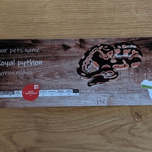 May include: A brown and white wooden sign with the text "Your pet's name Royal Python Python regius" and a picture of a brown and black snake. The sign also includes a red circle with the text "Near Threatened" and a green outline of the continent of Africa.