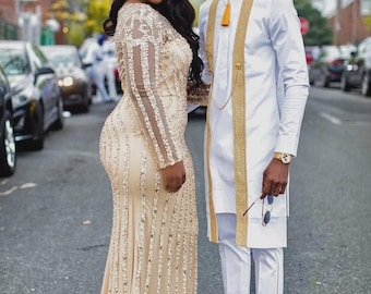 May include: A couple in formal wear pose on a street. The woman wears a long, gold sequined gown with sheer sleeves. The man is in a white outfit with gold trim and accessories. Cars are parked in the background.