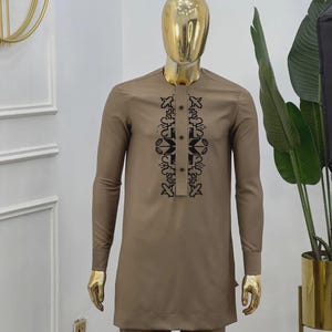 May include: A brown two-piece outfit with long sleeves and a mandarin collar. The shirt has a black geometric design down the front and is buttoned. The outfit is displayed on a gold mannequin.