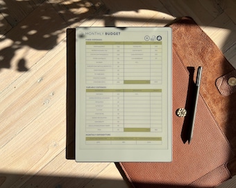 May include: A beige monthly budget planner with a black stylus on a brown leather surface. The planner has sections for fixed expenses, variable expenses, and monthly expenditure.