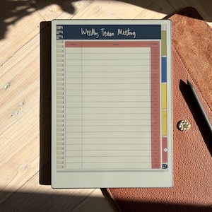 May include: A white weekly team meeting planner with a blue header that says "Weekly Team Meeting". The planner has a red, yellow, and blue color scheme and is on a brown leather surface.
