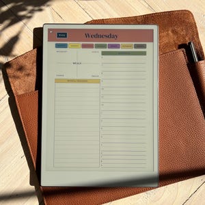May include: A gray e-reader with a white screen displaying a weekly planner template. The template is for Wednesday and includes sections for breakfast, lunch, dinner, snacks, and notes and thoughts. The screen also includes a schedule section with a list of times from 9 am to 8 pm.