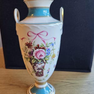 May include: A white porcelain vase with two side handles, featuring a central floral bouquet in a basket tied with a pink ribbon. The neck and base are accented with turquoise bands and gold trim, while the body displays a colourful arrangement of roses and wildflowers.
