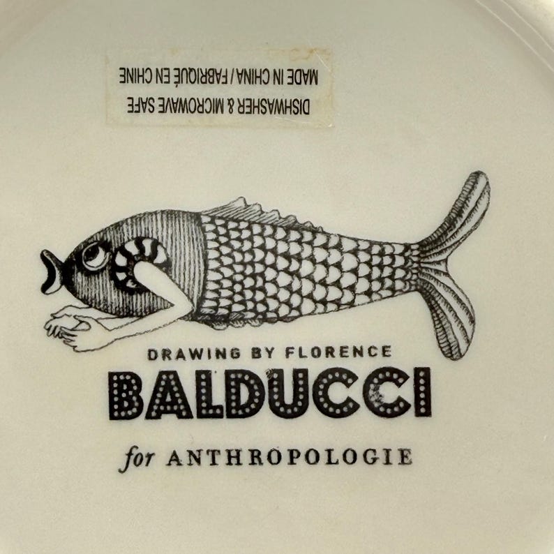 May include: A white ceramic dish with a black and white illustration of a fish. The fish design is by Florence Balducci for Anthropologie. Dishwasher and microwave safe. Made in China.