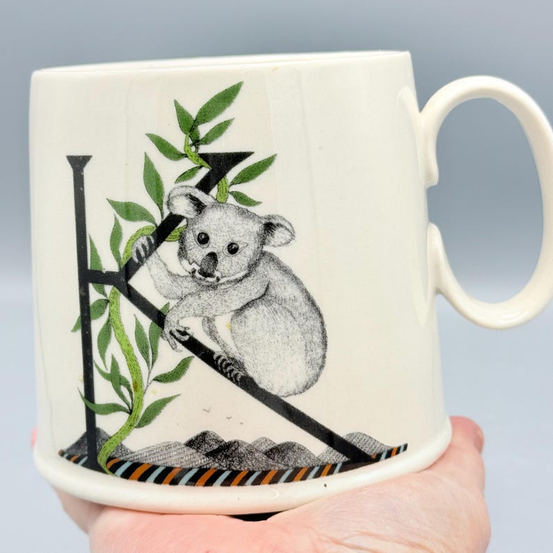 May include: Cream-colored ceramic mug featuring a black and white illustration of a koala clinging to a branch forming the letter "K". Green leaves and a mountain range are also depicted. The mug has a large, curved handle.