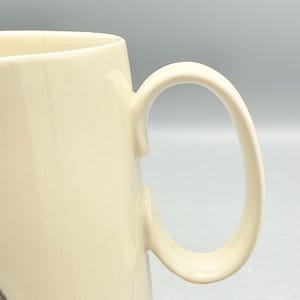 May include: Cream-colored ceramic mug with a curved handle. The mug has a smooth, glossy finish and a simple, classic design. The handle is large and easy to grip, making it suitable for hot or cold beverages.